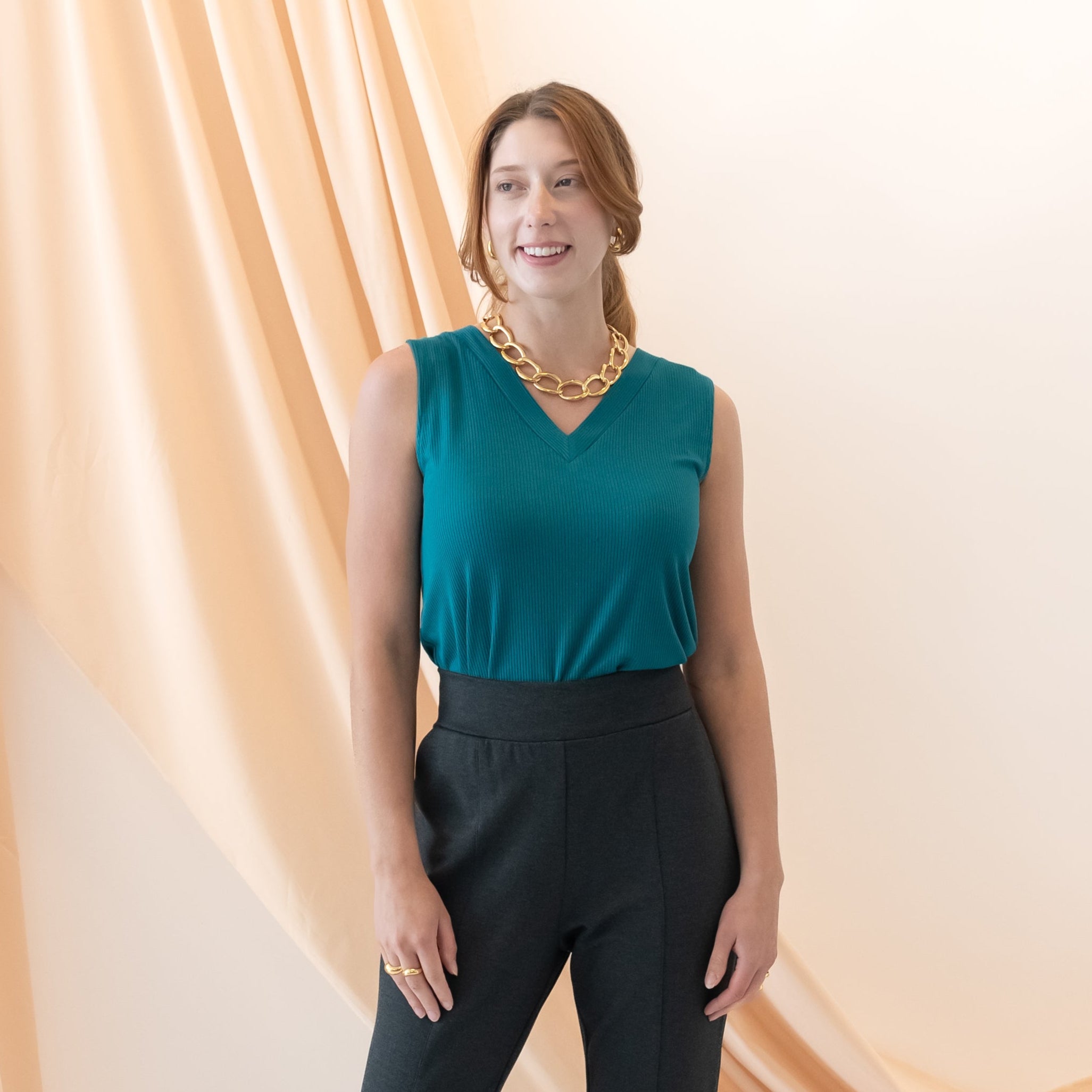 Woman wearing a teal sleeveless top and black pants standing against a beige curtain.