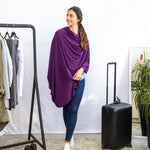 Woman wearing a purple poncho standing next to a black suitcase in a room with a clothing rack and plant.